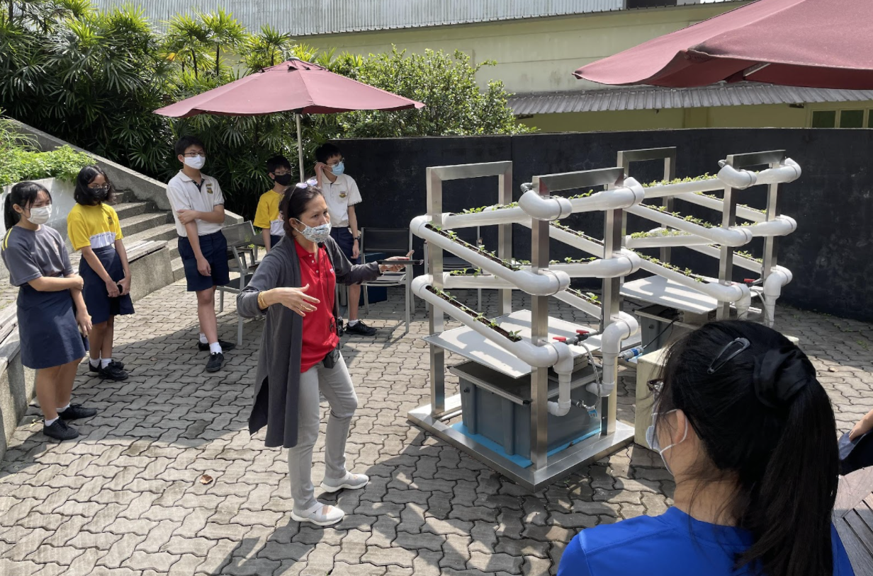 A masked woman gestures toward a hydroponic PVC pipe growing system while students in uniforms watch outdoors.